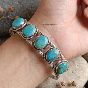 May include: A silver cuff bracelet featuring five oval turquoise stones set in twisted rope bezels. The metal band displays etched detailing between each stone. The text 925SilverJewelry26 is visible on the surface of the bracelet.