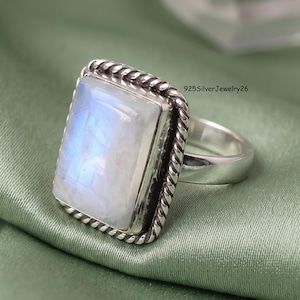 May include: A sterling silver ring featuring a large, rectangular, cabochon-cut moonstone with a blue iridescent sheen. The stone is set in a bezel with a twisted rope-style border. The text 925SilverJewelry26 is visible near the ring setting.