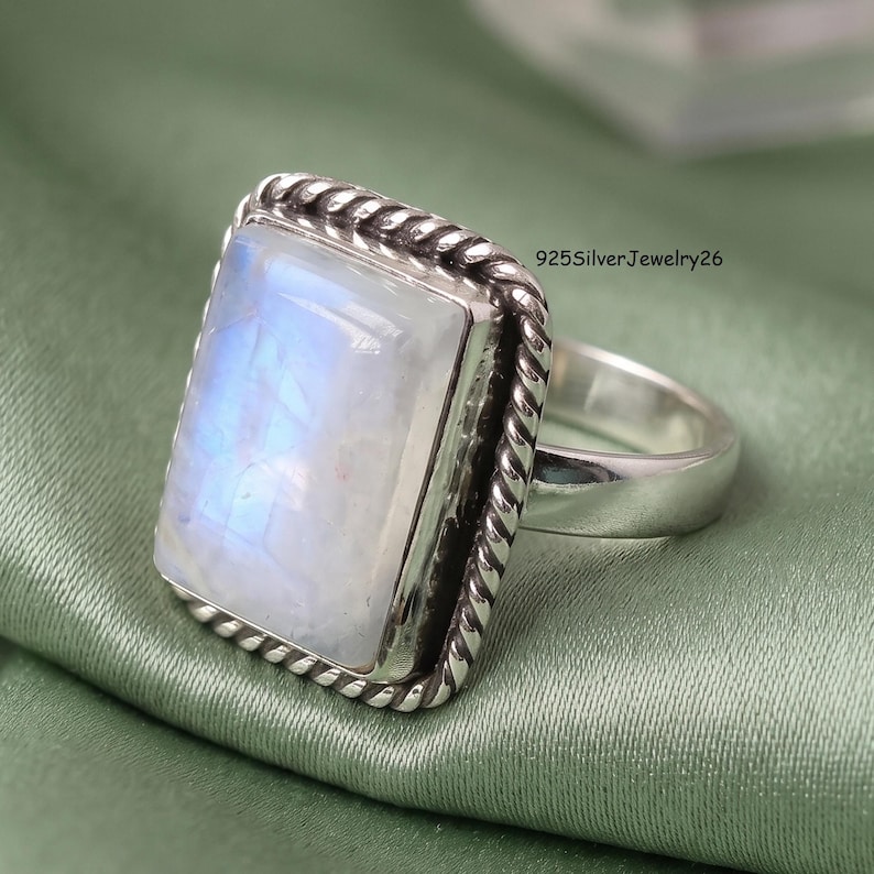 May include: A sterling silver ring featuring a large, rectangular, cabochon-cut moonstone with a blue iridescent sheen. The stone is set in a bezel with a twisted rope-style border. The text 925SilverJewelry26 is visible near the ring setting.