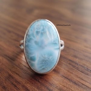 May include: An oval-shaped Larimar gemstone ring set in a polished silver band. The stone features a marbled pattern of light blue and white tones with a smooth, reflective surface. The text 925SilverJewelry26 is visible in the upper right corner.