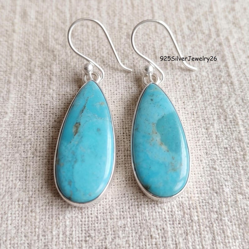 May include: A pair of teardrop-shaped turquoise earrings set in silver with curved ear wires. Each stone features natural blue and green veining. The text 925SilverJewelry26 is visible in the upper right corner of the image.