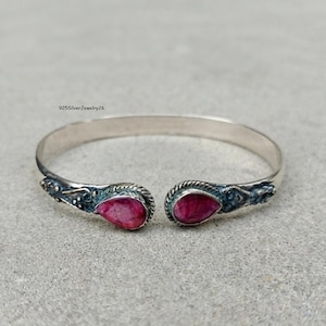 May include: A silver cuff bracelet featuring two teardrop-shaped red gemstones at the open ends. The metal band displays intricate oxidised detailing around the stone settings. The text 925SilverJewelry26 is visible above the bracelet.