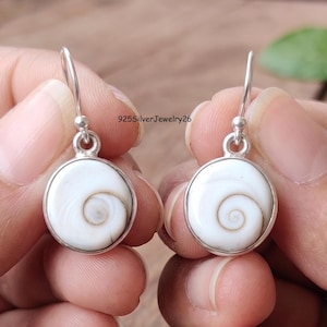 Op de afbeelding: Een paar hangende oorbellen met ronde witte Shiva-oog schelp cabochons gezet in gepolijste zilveren randen met zilveren haken. De tekst 925SilverJewelry26 is zichtbaar tussen de oorbellen. De schelpen tonen een natuurlijk spiraalpatroon.