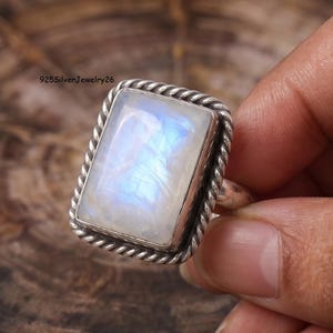 May include: A rectangular moonstone ring set in a silver band with a twisted rope-style border. The gemstone displays a translucent white appearance with iridescent blue flashes. The text 925SilverJewelry26 is visible in the upper left corner.