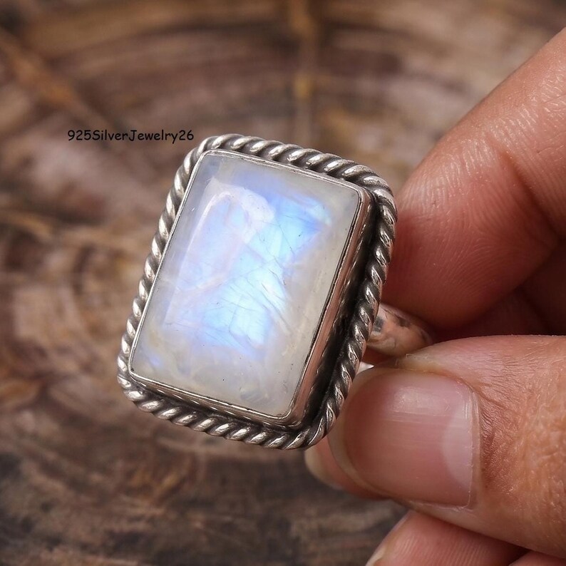 May include: A rectangular moonstone ring set in a silver band with a twisted rope-style border. The gemstone displays a translucent white appearance with iridescent blue flashes. The text 925SilverJewelry26 is visible in the upper left corner.