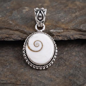 Puede incluir: Un colgante de plata con una concha de ojo de Shiva blanca y redonda con un patrón de espiral marrón natural. La concha está engastada en un marco de plata trenzada con un asa decorativa y ornamentada en la parte superior. El texto 925SilverJewelry26 es visible.