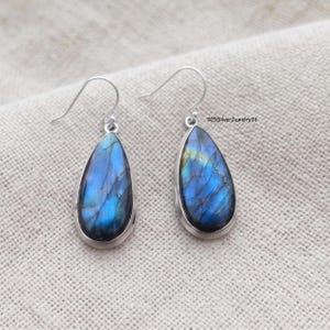 Puede incluir: Un par de pendientes de labradorita en forma de lágrima engastados en plata con ganchos. Las piedras muestran destellos iridiscentes azules y dorados en sus superficies. El texto 925SilverJewelry26 es visible cerca del pendiente derecho.
