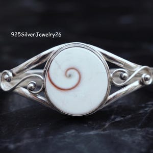 Puede incluir: Un brazalete de plata con una concha de ojo de Shiva blanca circular en el centro, con un patrón de espiral marrón. La banda de metal tiene detalles de alambre en espiral decorativos a ambos lados. El texto 925SilverJewelry26 es visible.