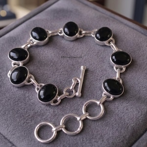 Op de afbeelding: Een zilveren armband met zeven ovale zwarte stenen in gepolijste zilveren zettingen, verbonden door zilveren schakels. De armband heeft een staafsluiting en een korte verlengketting met drie ronde schakels. De tekst 925SilverJewelry26 is zichtbaar.