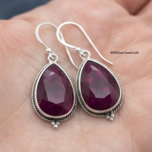 May include: A pair of teardrop-shaped earrings featuring faceted deep magenta stones set in silver frames with a twisted rope border and silver hook wires. The text 925SilverJewelry26 is visible in the upper right corner of the image.