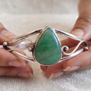 May include: A silver cuff bracelet featuring a central teardrop-shaped green stone. The metal band has a polished finish with decorative swirling wire details on both sides of the stone setting, creating an open, symmetrical design.