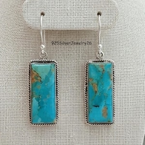 Puede incluir: Un par de pendientes colgantes rectangulares con piedras de turquesa pulidas con patrones de matriz marrón, engastados en marcos de plata con bordes de cuerda trenzada y ganchos de plata. El texto 925SilverJewelry26 es visible entre los pendientes.