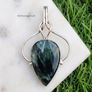 Puede incluir: Un colgante en forma de lágrima con una piedra preciosa de serafinita verde oscuro con inclusiones blancas plumosas, engastado en un marco de plata de ley pulida con un pasador decorativo. El texto 925SilverJewelry26 es visible en la parte superior.