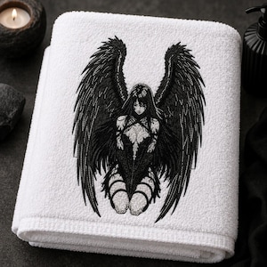 May include: A white bath towel featuring a detailed black embroidered graphic of a winged figure with long hair and intricate clothing. The embroidery depicts a central character with large, feathered wings spread wide, rendered in high-contrast black thread.