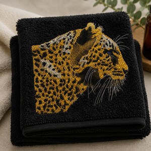 May include: A black hand towel featuring a detailed embroidered design of a leopard head in profile. The embroidery uses gold, yellow, and black thread to depict the animal's spotted fur, whiskers, and facial features against the dark fabric.