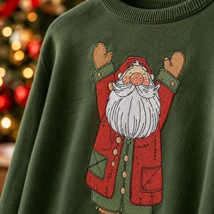 May include: A dark green long-sleeve sweatshirt featuring a large embroidered graphic of a bearded Santa Claus figure with arms raised. The figure wears a red coat, green trousers, and tan boots, with detailed stitching throughout the design.
