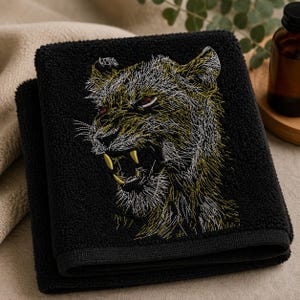 May include: A black hand towel featuring an embroidered design of a snarling big cat. The embroidery uses white and gold thread to depict the animal's fur, with prominent gold fangs and dark red eyes visible against the textured black fabric.