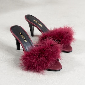 Pode incluir: Mules bordô de salto alto com acabamento em penas fofas. Os saltos pretos e as palmilhas apresentam a inscrição dourada "ROSALIND". Os sapatos são exibidos em uma superfície cinza claro. Os saltos medem cerca de 7,6 cm.
