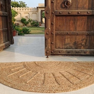 May include: A half-circle jute doormat with a sunburst design lies in front of a large, dark wooden door. The mat is made of woven natural fibers. Potted plants with orange flowers flank the doorway.