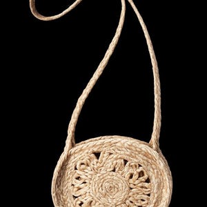 Puede incluir: Un bolso de hombro redondo de paja trenzada de color beige con un patrón circular texturizado en la parte frontal. El bolso incluye una correa larga de paja trenzada unida a los lados superiores, creando un asa continua para llevar al hombro.