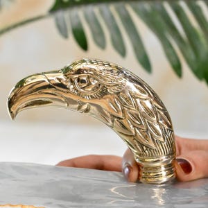 May include: A gold-colored eagle head sculpture, featuring detailed feather and eye carvings. The beak is curved and pointed. The sculpture is held by a person's hand, with a blurred background of green leaves.