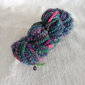 May include: A colorful skein of yarn with a mix of blue, purple, pink, and green hues. The yarn has a textured appearance with a beaded accent and a dangling purple charm. The yarn is displayed on a white, textured surface.