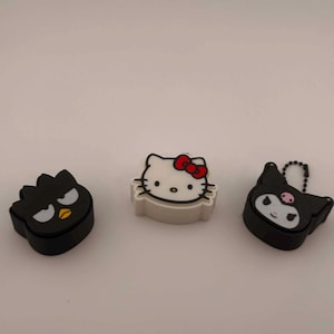 May include: Three small character-shaped silicone covers featuring Badtz-Maru, Hello Kitty, and Kuromi. Each item has a distinct facial expression and color scheme, including black, white, and red accents, designed as decorative accessories.