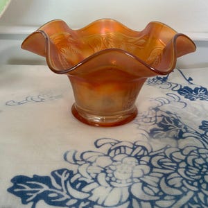 May include: An iridescent orange glass bowl with a ruffled rim. The bowl has a wide, shallow shape and a pedestal base. The glass has a textured surface with a wheat pattern. The bowl is sitting on a white and blue floral patterned cloth.