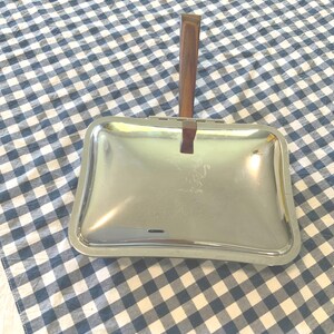 May include: A rectangular silver-toned metal crumb sweeper featuring a polished finish and a brown wood-grain handle. The top surface displays a faint etched graphic of a small figure, and the device includes a hinged mechanism for operation.