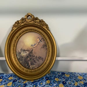 May include: An oval wall art piece featuring a gold-colored frame with ornate floral detailing at the top. The print inside depicts two small birds perched on a branch near a bird's nest, set against a soft, glowing yellow sunlit background.