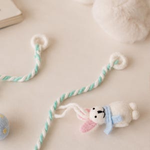Puede incluir: Un adorno de fieltro blanco con forma de conejo, con orejas rosadas y una bufanda azul claro, unido a un cord&oacute;n trenzado de color blanco y verde menta. El cord&oacute;n presenta un peque&ntilde;o lazo circular en el extremo. El conejo tiene ojos negros.