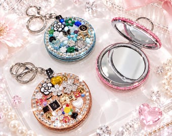 May include: Two round compact mirrors with keychains, one blue and one rose gold, decorated with various charms including flowers, butterflies, and perfume bottles. A third open rose gold compact mirror shows a reflective surface.