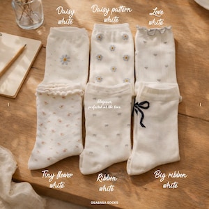 May include: A collection of white socks with various designs, including daisies, tiny flowers, and ribbons. The socks are displayed on a wooden surface, with text labels indicating the style of each pair. The text "Elegance, perfected at the toes" is also visible.