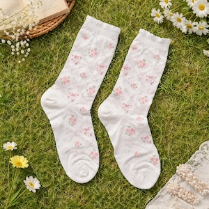 May include: A pair of white socks with a floral pattern. The socks feature a repeating design of pink roses and bows within a diamond grid. The socks are displayed on a bed of green grass, with daisies and a book in the background.
