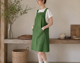 May include: A forest green sleeveless apron dress featuring a square neckline, wide shoulder straps, and two large front patch pockets. The dress is worn over a plain white short-sleeved t-shirt and paired with white socks, reaching above the knees.