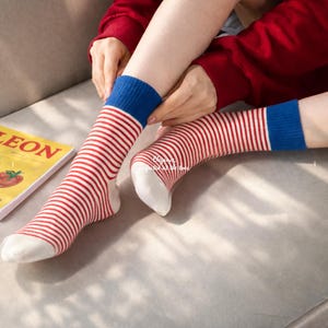May include: A pair of striped socks with red and white stripes, a blue cuff, and a white toe. The socks are being adjusted by a person. A book with the word "LEON" is visible in the background.