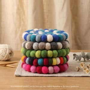 以下が含まれることがあります： 青、緑、ピンクなど、さまざまな色の手作りのフェルトボールコースターのスタック。 コースターは、ニュートラルカラーのリネンの表面に配置されています。 下部のテキストには、「これは手作りの製品であり、色は製造時期によって若干異なる場合があります」と書かれています。