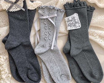 May include: Three pairs of ribbed socks in shades of charcoal and light gray. The first pair features a cream lace cuff with a gray ribbon bow. The second pair has a light gray ribbon bow and vertical ruffles. The third pair includes a white lace patch.
