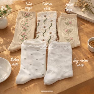 Puede incluir: Una variedad de calcetines beige y blancos con diseños florales y de lazo. Los calcetines incluyen bordados de tulipanes, cosmos y corazones, así como calcetines con pequeños patrones de lazo. El texto "OSABASA SOCKS" es visible.