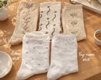 May include: An assortment of beige and white socks with floral and bow designs. The socks include embroidered tulips, cosmos, and hearts, as well as socks with tiny ribbon patterns. The text "OSABASA SOCKS" is visible.
