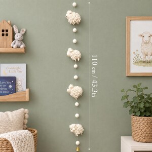Puede incluir: Un colgante de pared vertical con cuatro figuras de ovejas texturizadas de color crema intercaladas con peque&ntilde;as bolas de fieltro blancas en un hilo fino. La longitud total es de 110 cm y termina con una peque&ntilde;a campana dorada en la parte inferior.