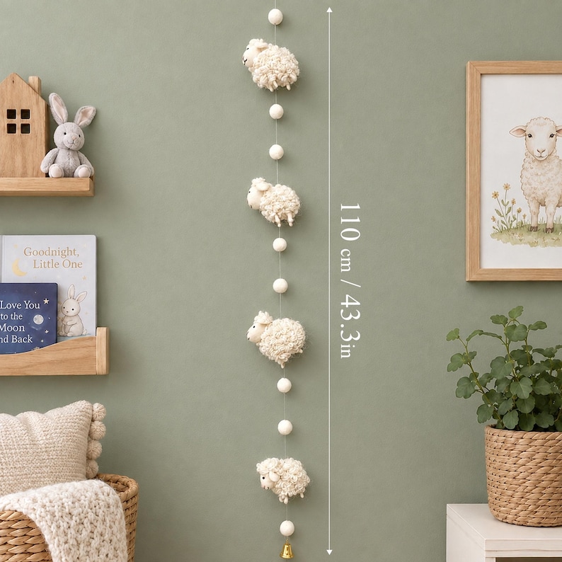 Puede incluir: Un colgante de pared vertical con cuatro figuras de ovejas texturizadas de color crema intercaladas con peque&ntilde;as bolas de fieltro blancas en un hilo fino. La longitud total es de 110 cm y termina con una peque&ntilde;a campana dorada en la parte inferior.