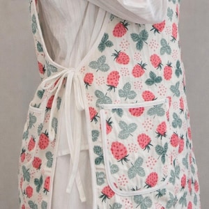 May include: A white apron with a repeating pattern of pink strawberries and green leaves. The apron has two pockets and white ties at the waist. The apron is made of a lightweight fabric.