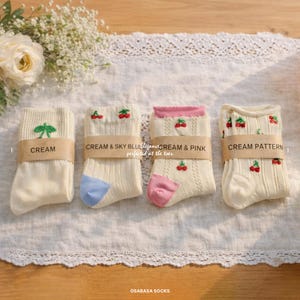 May include: Four pairs of cream-colored socks with various designs. One pair has a green leaf, another has cherries with blue accents, a third has cherries with pink accents, and the last has a cherry pattern. Each pair is wrapped with a brown band.