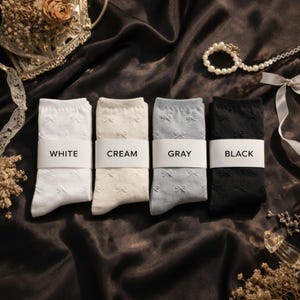 Puede incluir: Cuatro pares de calcetines en blanco, crema, gris y negro, cada uno con un patrón de lazo. Los calcetines están cuidadosamente doblados y etiquetados con sus respectivos colores. El fondo es una tela oscura y texturizada.