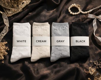 May include: Four pairs of socks in white, cream, gray, and black, each with a bow pattern. The socks are neatly folded and labeled with their respective colors. The background is a dark, textured fabric.