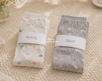 May include: Two pairs of socks, one white and one gray, are displayed. Each pair is folded and wrapped with a white band labeled "WHITE" and "GRAY". The socks feature a floral pattern and the text "Elegance, perfected at the toes."