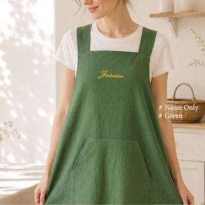 May include: A forest green apron featuring wide shoulder straps and a large front pocket. The name Jessica is embroidered in yellow cursive script on the chest. The apron is worn over a plain white short-sleeved t-shirt.