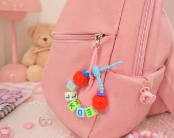 May include: A light pink backpack featuring a zipper pull adorned with a circular charm made of colorful plastic beads. The charm includes red spherical beads, light blue accents, and square white beads with green and blue letters.