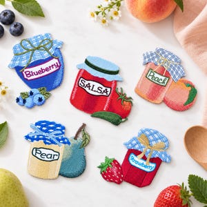 May include: Five embroidered fabric patches shaped like jars of preserves and salsa. The patches feature blue, red, peach, yellow, and red jars with gingham lids, labeled Blueberry, Salsa, Peach, Pear, and Strawberry, with matching fruit accents.